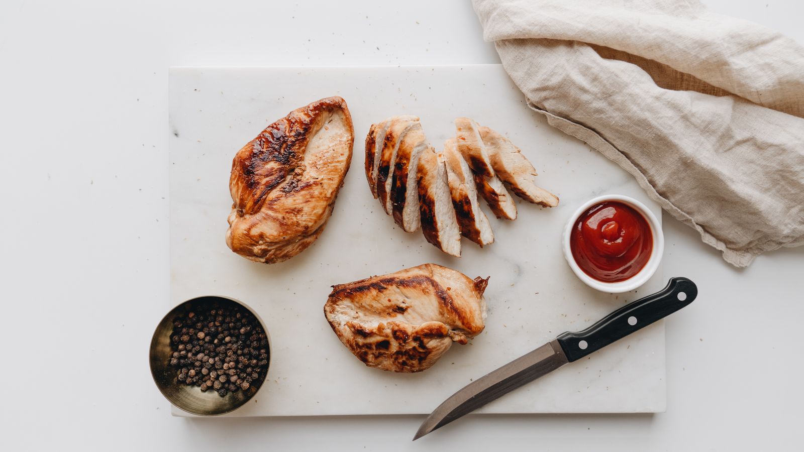 Grilled chicken breasts, one whole and one sliced, arranged on a marble board with dipping sauce and peppercorns.