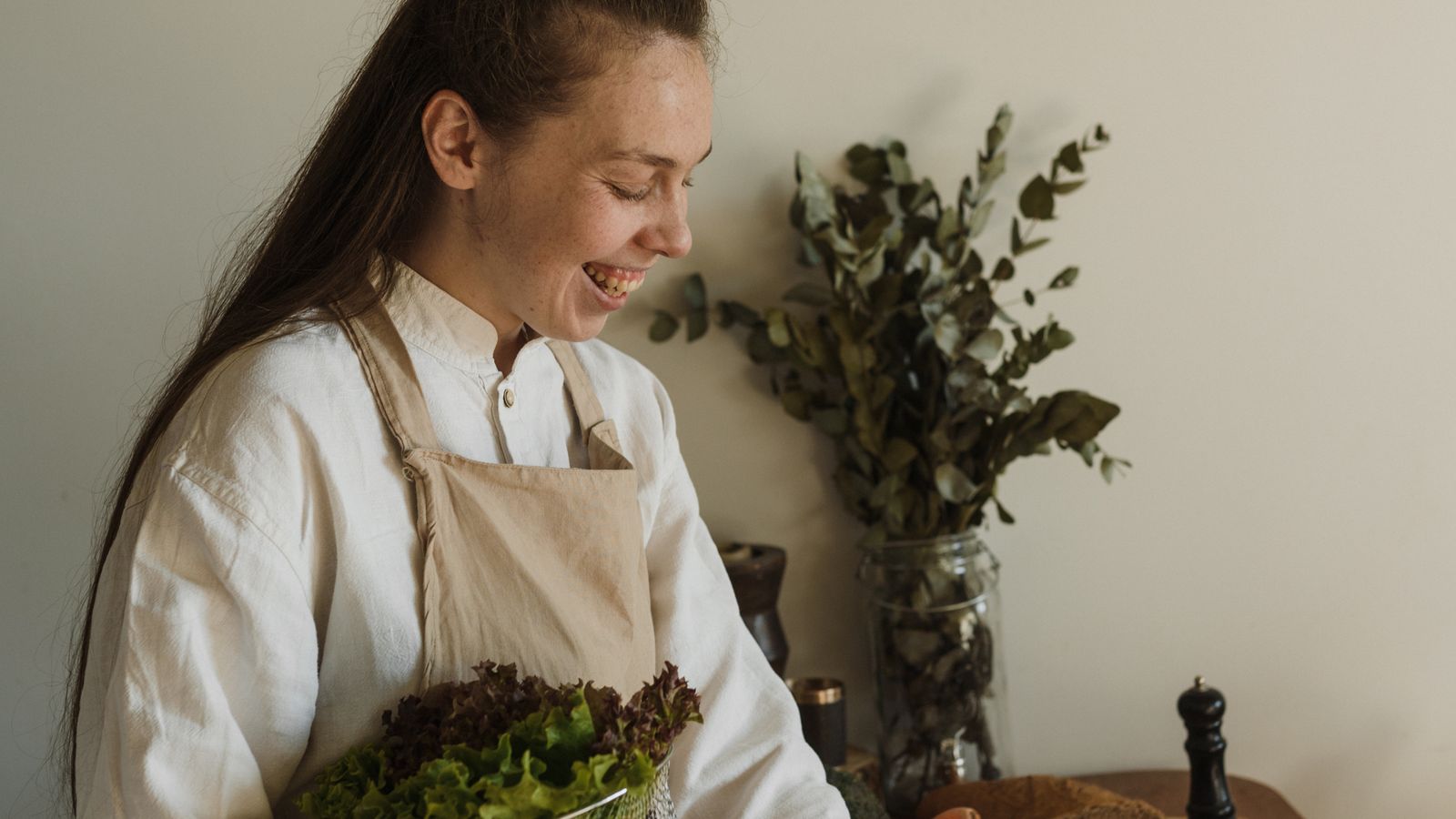 A smiling woman in an apron holding a bowl of fresh salad greens while preparing a healthy meal in her kitchen.